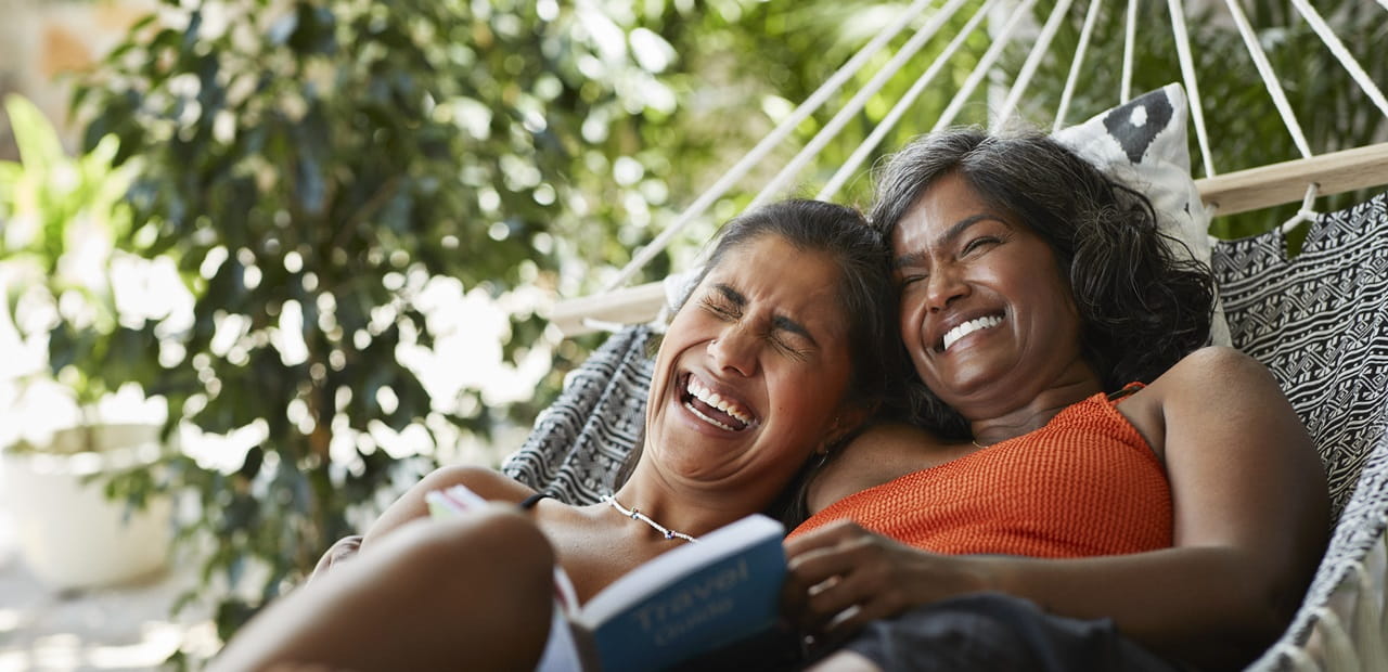 A mother and daughter lie together in a hammock surrounded by lush greenery, with the mother wearing an orange top and holding a book, while the daughter laughs while resting her head on her mother's shoulder.