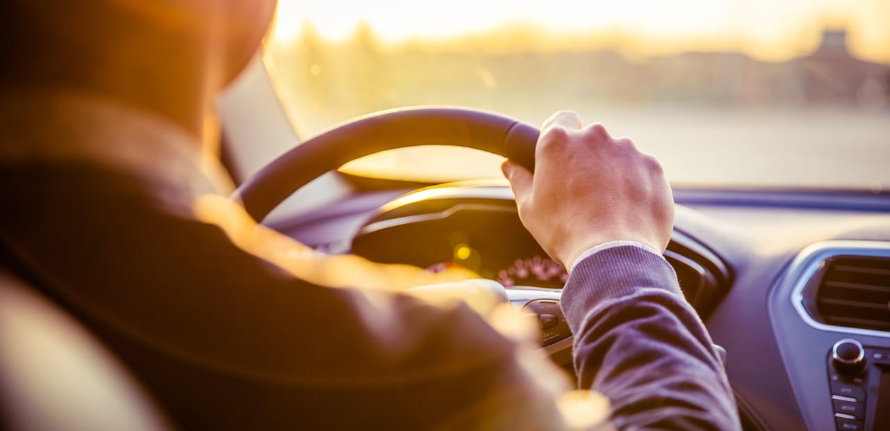 A person drives a car during sunset, viewed from the back seat with sunlight streaming through the windshield and the dashboard softly illuminated.